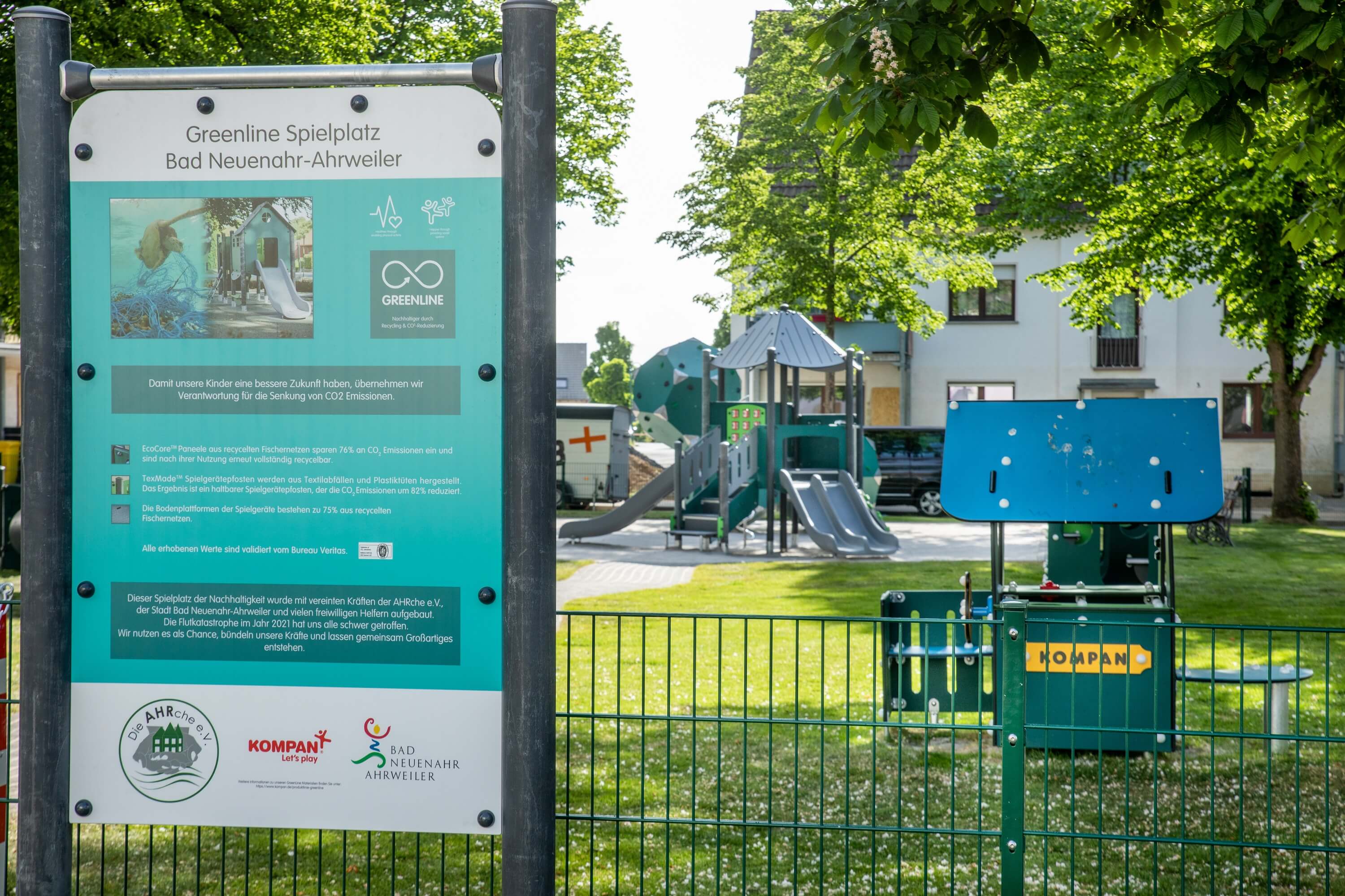 Information board for “Greenline Spielplatz” in front of a fenced, green playground with slides and climbing structures in a park