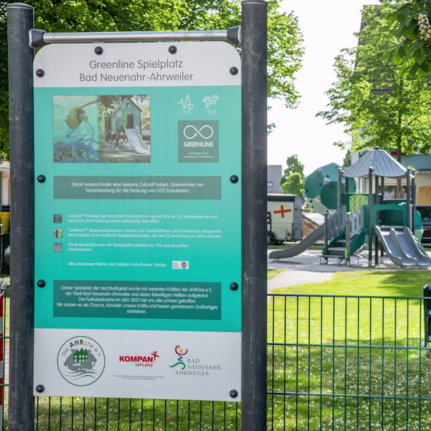 Information board for “Greenline Spielplatz” in front of a fenced, green playground with slides and climbing structures in a park
