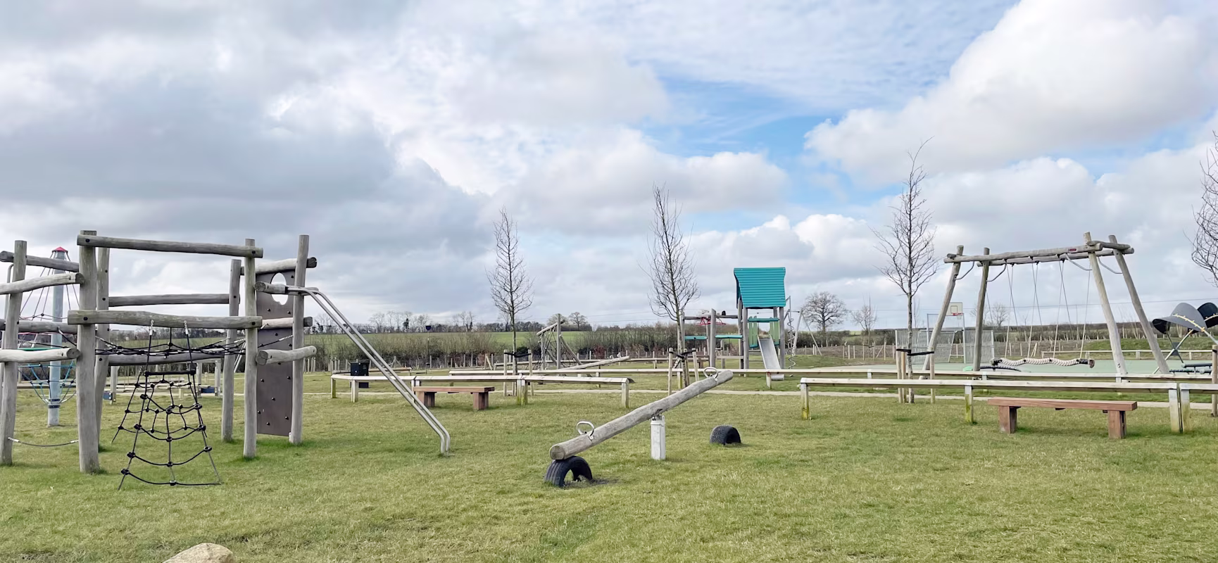 Empty outdoor playground with wooden climbing frame, swings and seesaw on green grass under a cloudy blue sky.