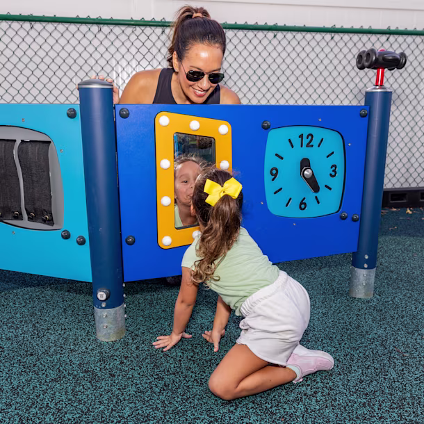 a preschool age girl making a silly face in a play panel mirror while her mother lovingly overlooks her