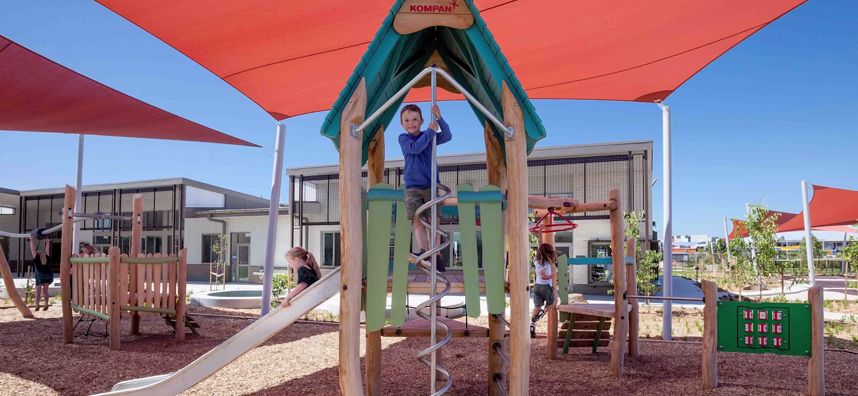 Children play on a shaded outdoor playground with slides, climbing poles and wooden structures under large orange sun canopies.