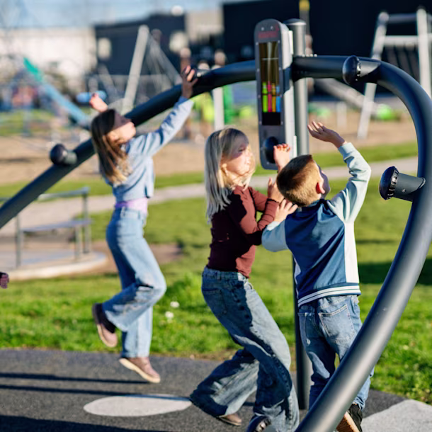 Children play on a modern grey climbing and game structure in a sunny playground with grass and swings in the background.