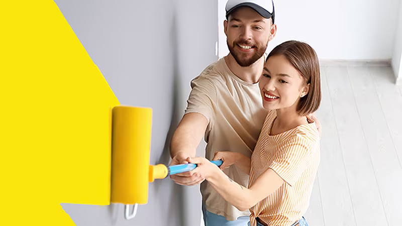 Koppel schildert samen een muur in geel met een verfroller; ze lachen en genieten van het moment. | Un couple peint ensemble un mur en jaune avec un rouleau, tout en souriant et profitant du moment.