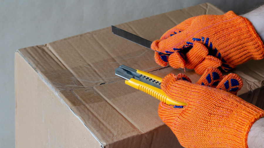 Een man met orange werkhandschoenen vervangt het mesje van een breekmes boven een kartonnen doos | Un homme portant des gants de travail orange replace la lame d'un couteau à briser au-dessus d'une boîte en carton