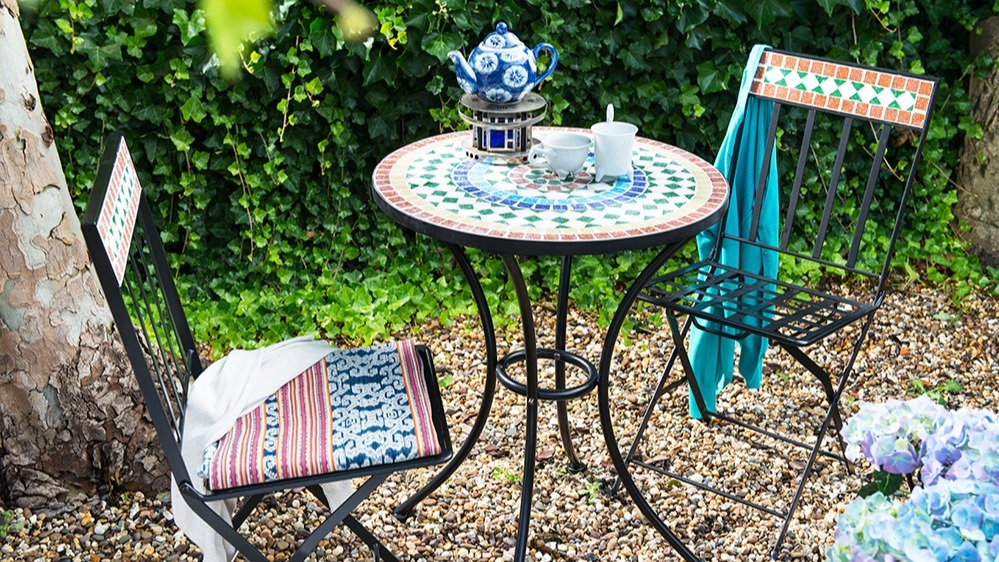 Twee mozaïeken en een mozaïek tafeltje staan op het grind in de tuin met kopjes thee erop | Deux mosaïques et une petite table en mosaïque sont posées sur le gravier du jardin avec des tasses de thé dessus.