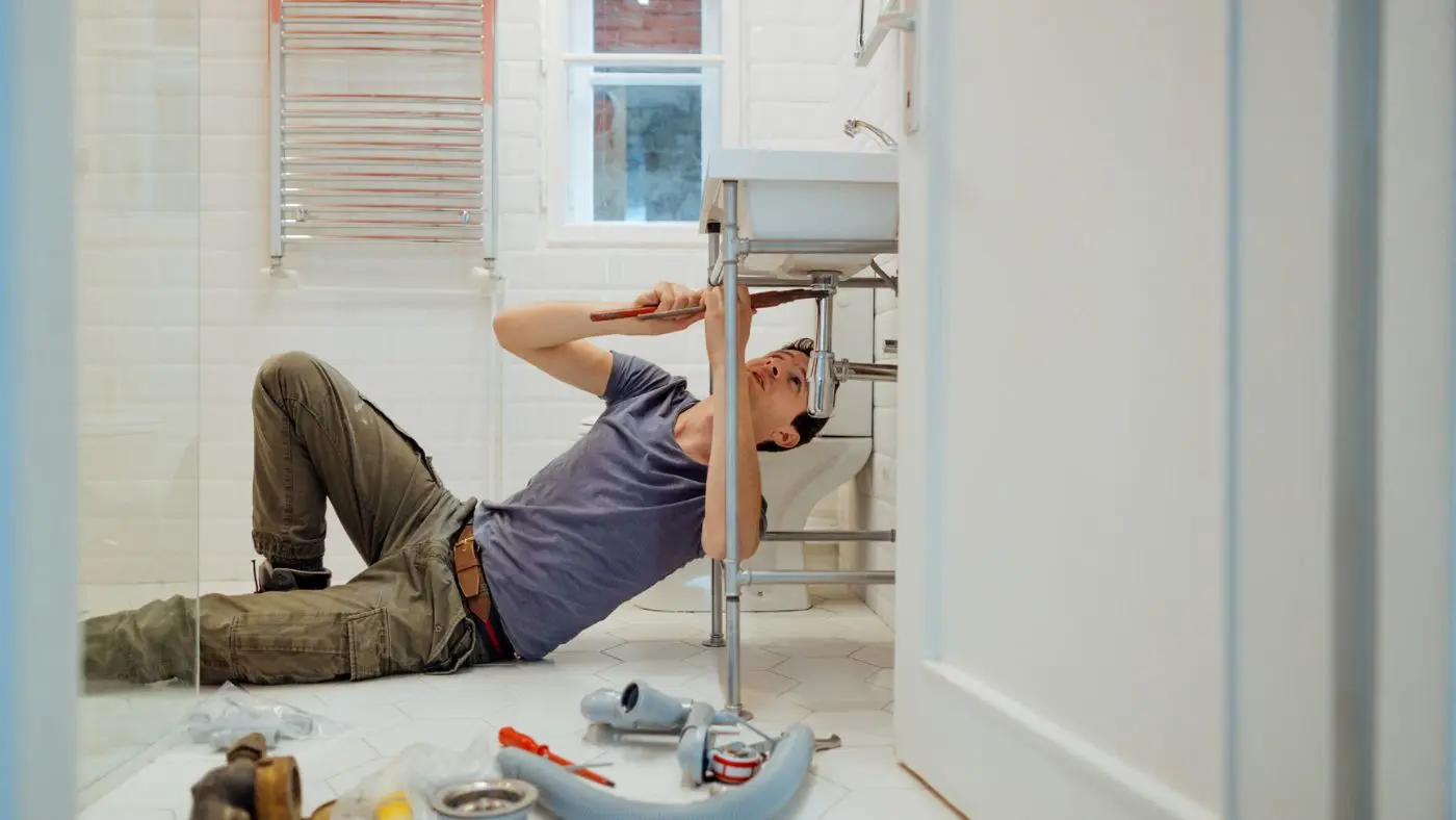 Doe-het-zelver repareert een wastafel in een badkamer. Diverse gereedschappen liggen verspreid op de grond | Un bricoleur répare un lavabo dans une salle de bain. Divers outils sont éparpillés sur le sol