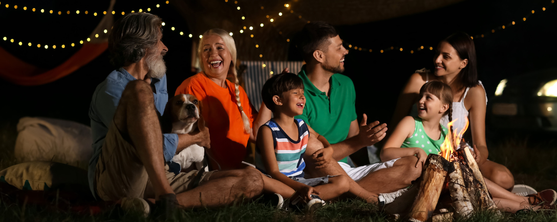 Een groep mensen 's avonds zittend op het gras voor een vuurtje in de tuin, met draadverlichting achteraan | Un groupe de personnes assises sur l'herbe la nuit devant un feu dans le jardin, avec des guirlandes lumineuses à l'arrière.