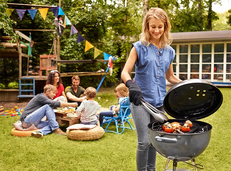 Een vrouw staat met een tang achter een barbecue | Une femme se tient debout avec des pinces derrière un barbecue