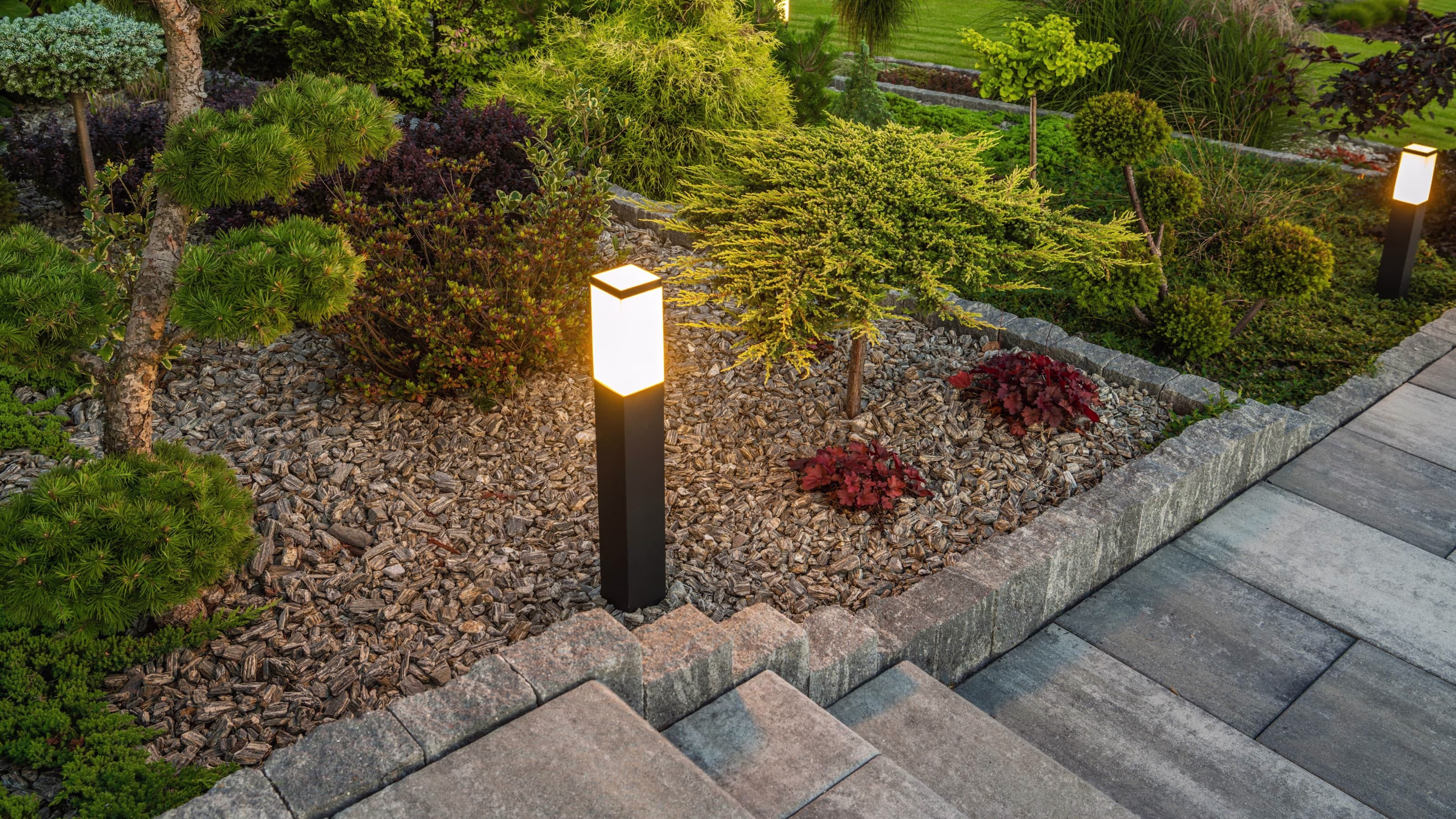 Moderne zwarte tuinpaalverlichting langs een tuinpad met siergrind, groenblijvende planten en boordstenen | Borne lumineuse de jardin noire moderne le long d’une allée avec paillis, plantes persistantes et bordures en pierre