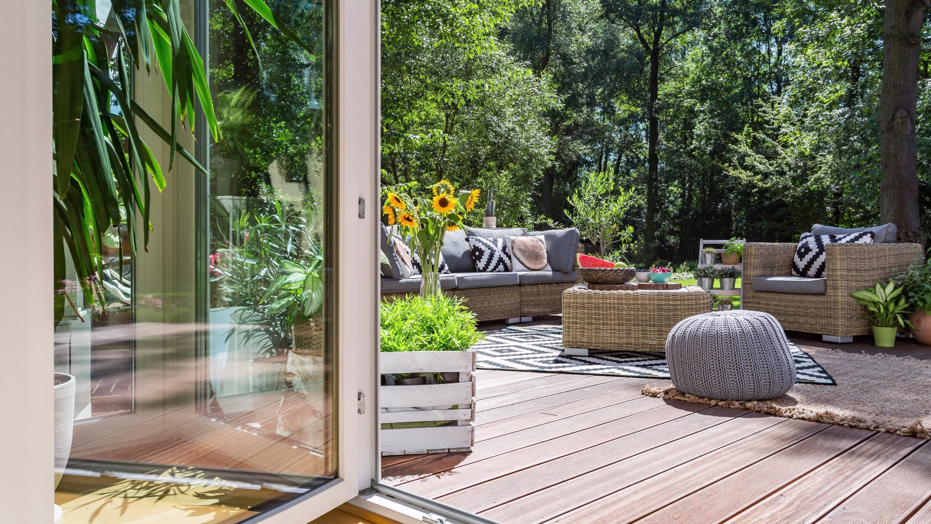 Gezellig houten terras met rotan tuinmeubelen, grijze kussens, zonnebloemen en groene tuin op de achtergrond | Terrasse en bois conviviale avec salon de jardin en rotin, coussins gris, tournesols et jardin verdoyant en arrière-plan