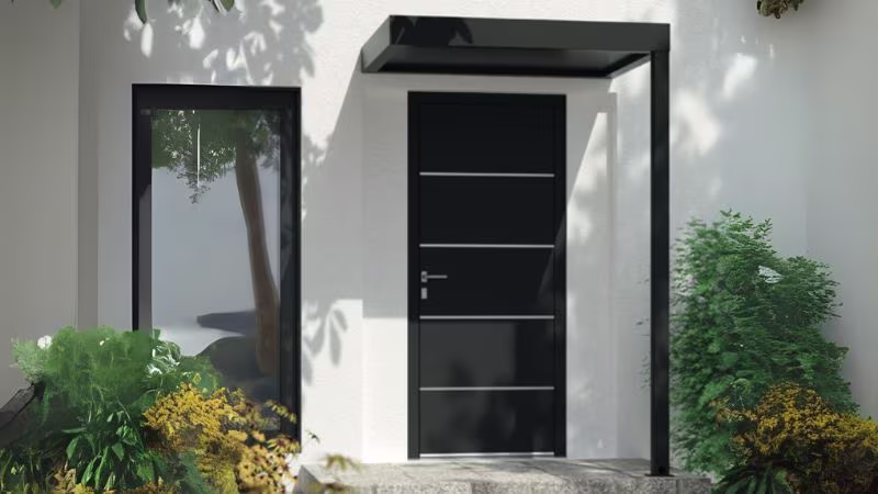 Moderne zwarte voordeur met strak luifelontwerp aan een witte gevel, omringd door planten. | Porte d'entrée noire moderne avec auvent épuré sur façade blanche, entourée de végétation.