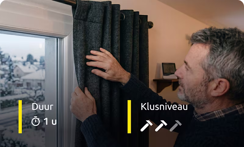 Man hangt thermische gordijnen op bij raam om warmte binnen te houden. | Homme installant des rideaux thermiques près d’une fenêtre pour conserver la chaleur.