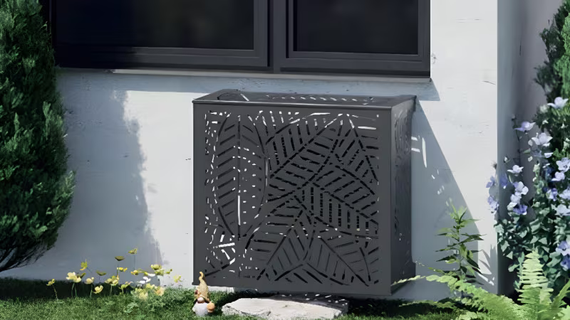 Decoratief rooster als airco-afscherming onder zwart raamkozijn tegen een witte gevel, omgeven door groen. | Grille décorative pour cacher la climatisation sous une fenêtre noire sur façade blanche, entourée de verdure.