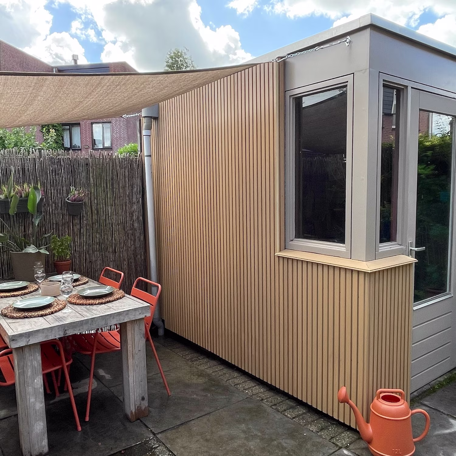 Schuur met een DIY lattenwand, tuintafel, oranje stoelen, oranje gieter en een schaduwdoek: praxis blog lattenwanden trends 2026