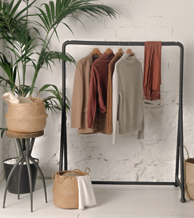 Metalen kleding rek met kleding, links een grote plant met daaronder een rieten wasmandje | Porte-linge en métal avec des vêtements, à gauche une grande plante avec un panier à linge en osier en dessous