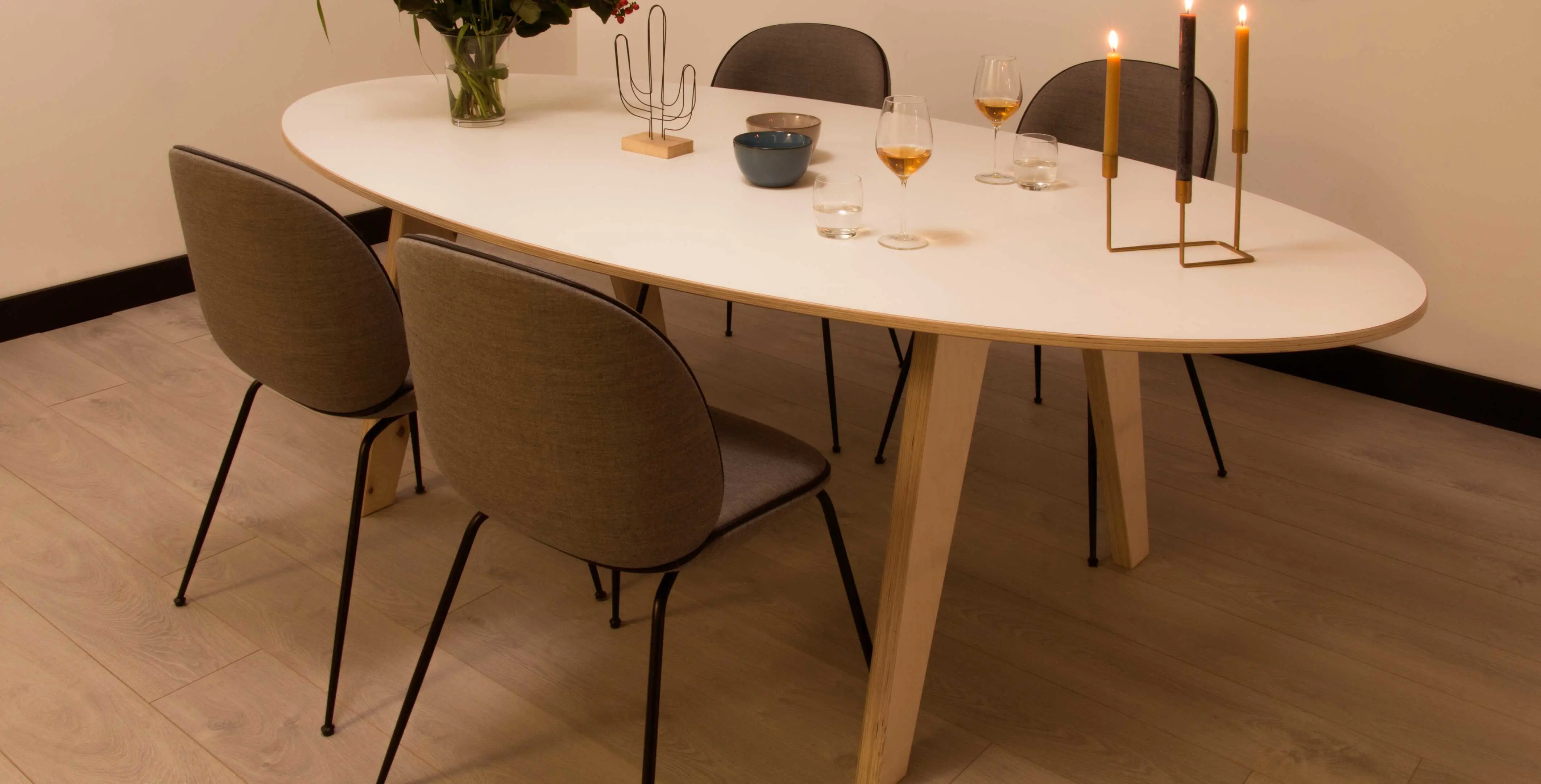 Ovale tafel met berken multiplex blad, omringd door grijze stoelen, gedekte tafel met kaarsen en glazen. | Table ovale avec plateau en contreplaqué de bouleau, entourée de chaises grises, dressée avec bougies et verres.