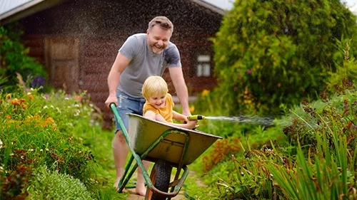 speelse man vervoert kind in kruiwagen en besproeien de tuin met water | Homme poussant sa fille dans une brouette qui arrose son jardin