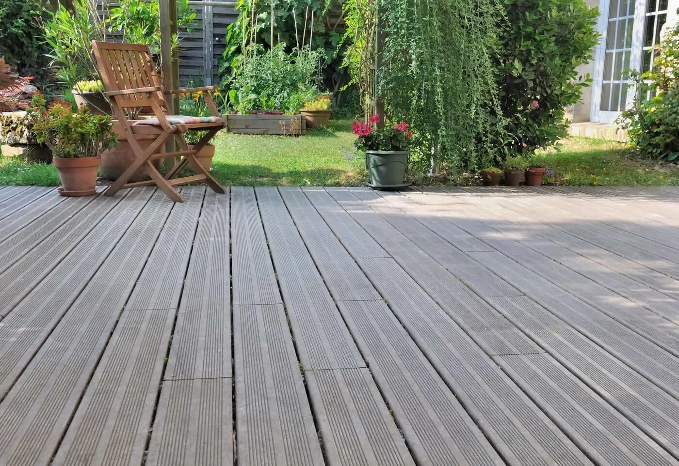 Een houten terras met een tuinstoel erop en erachter gras met planten | Une terrasse en bois avec une chaise de jardin et, derrière, de l'herbe avec des plantes