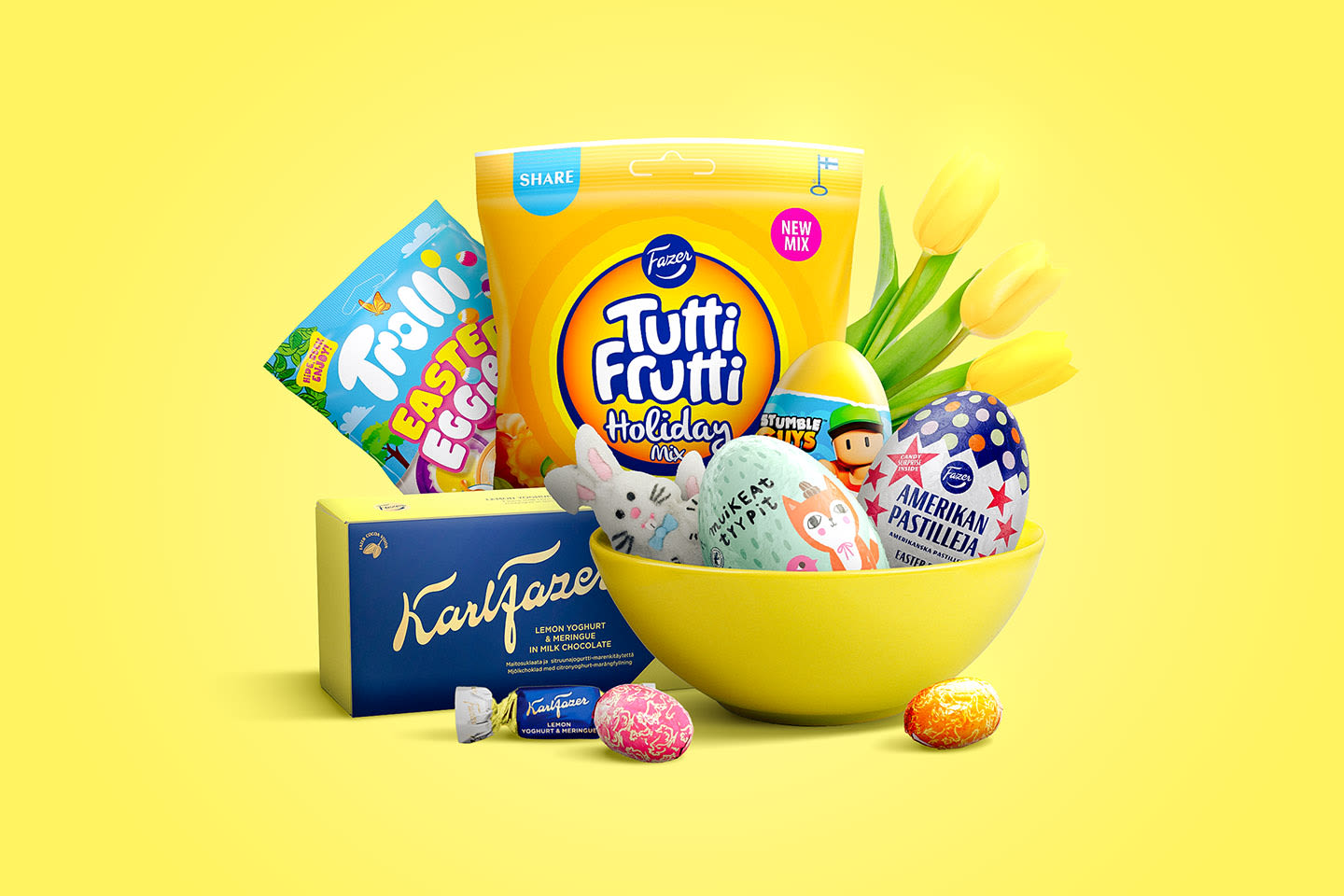 Keltaisessa kulhossa pääsiäisherkkuja: Fazerin makeisia, suklaamunia ja keltaisia tulppaaneja iloisessa asetelmassa.
