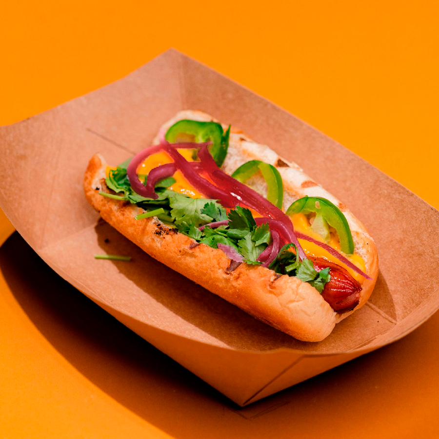 Mehevä hot dog rapeassa sämpylässä, täytteenä tuoretta korianteria, vihreitä jalapeno-siivuja ja punaisia sipulirenkaita.