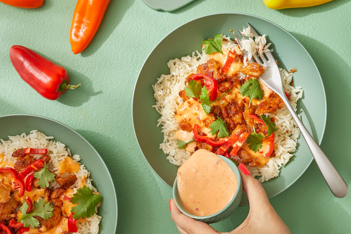 Mausteista riisiannosta vihreällä lautasella, jossa lihaa, paprikaa ja korianteria, käsi kaataa kastiketta päälle.