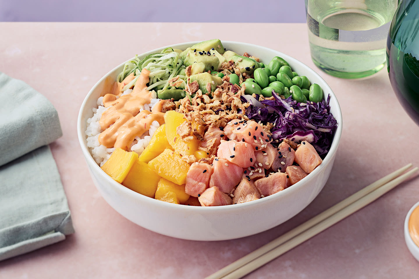 Värikäs poke bowl, jossa lohta, mangoa, avokadoa, edamamepapuja ja punakaalia riisin päällä valkoisessa kulhossa.