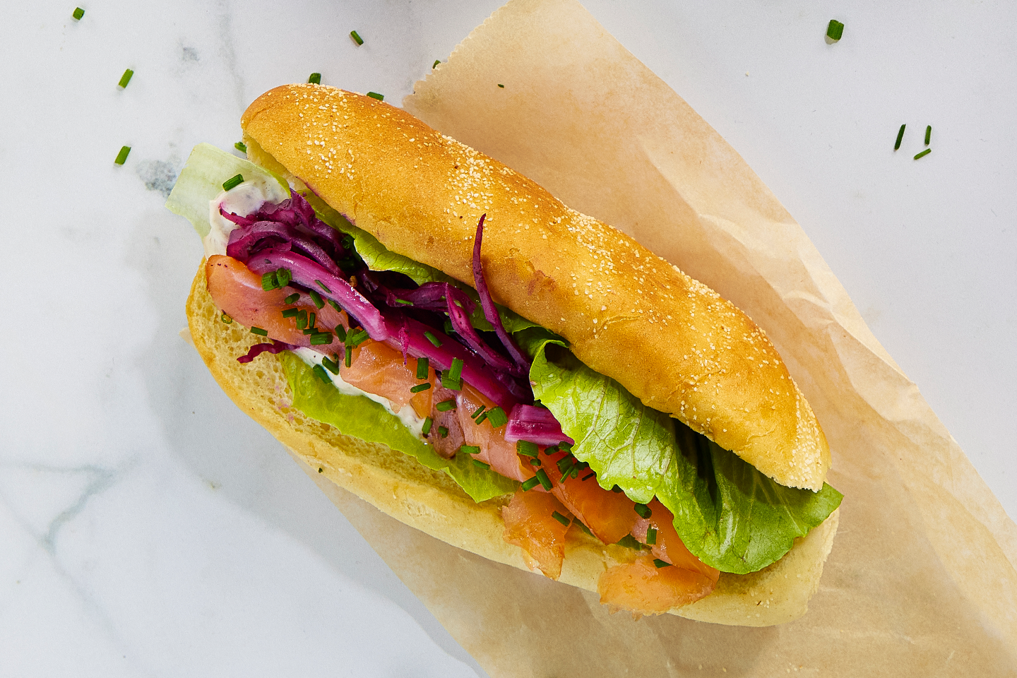 Hotdog-sämpylä täytettynä lohella, salaatilla, punakaalilla ja ruohosipulilla, aseteltuna ruskealle paperille.