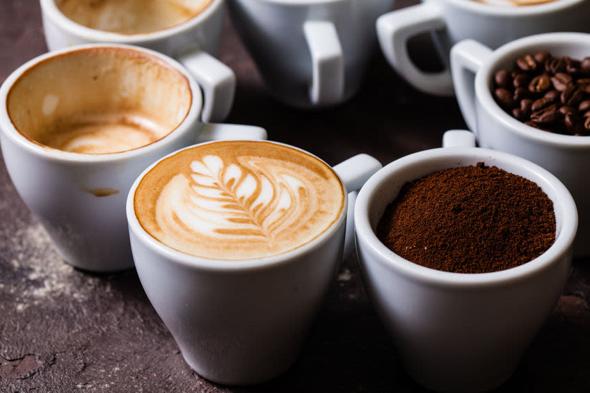 Useita kahvikuppeja, joissa yhdessä on Latte art -kukkakuvio, yhdessä kahvipapuja ja yhdessä hienoksi jauhettuja papuja