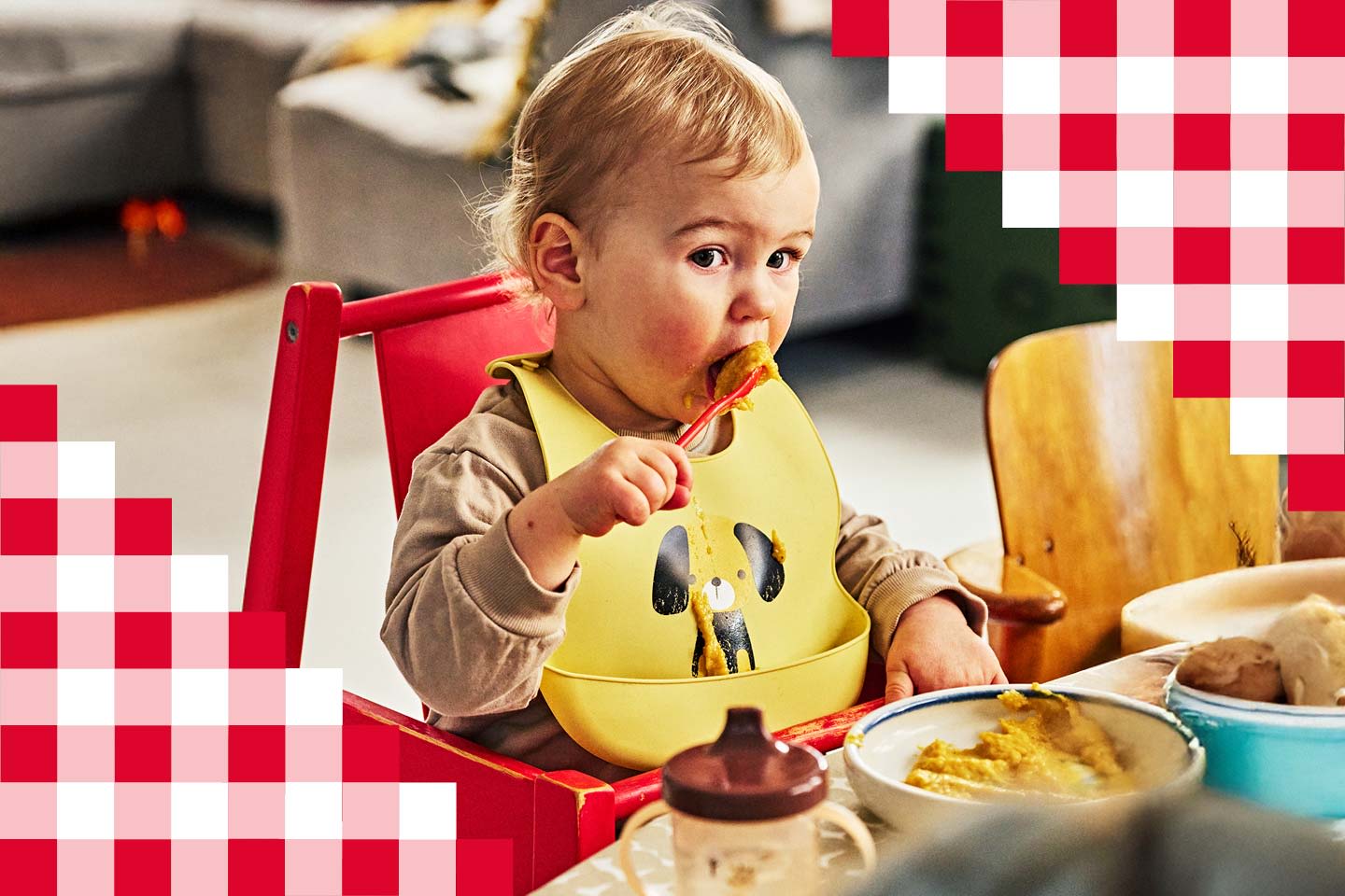 Pieni lapsi punaisessa syöttötuolissa nauttii ruokailusta keltainen koirakuvioinen ruokalappu yllään.