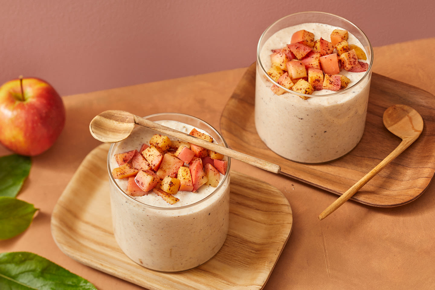 Kaksi lasillista kermaista jogurttijälkiruokaa puisilla alustoilla, päällä mausteisia omenanpaloja ja puiset lusikat vieressä.