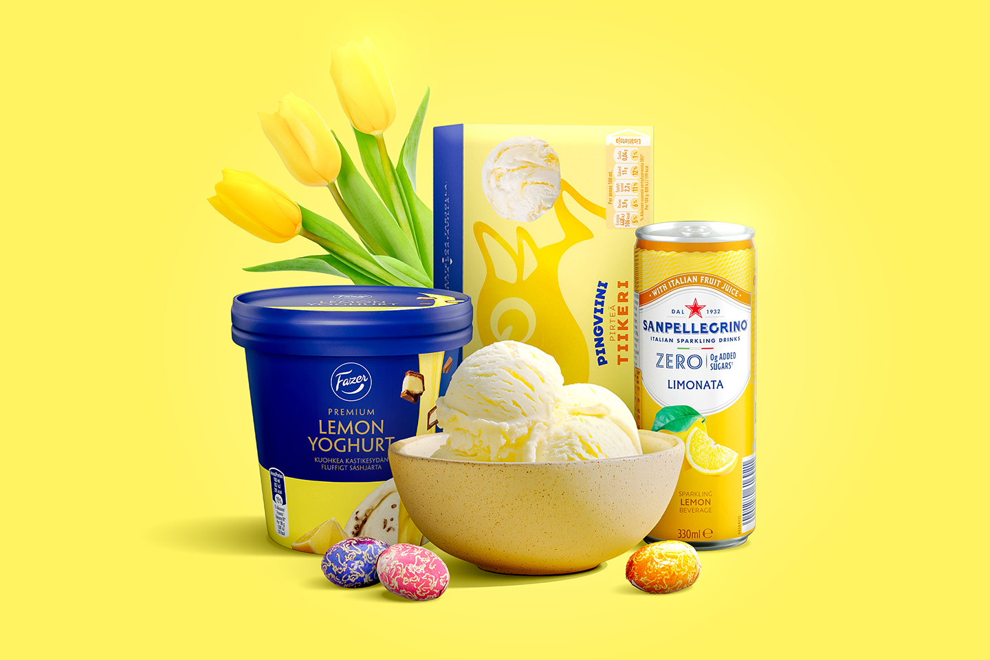 Keväinen keltainen asetelma, jossa jäätelöä kulhossa, Fazerin sitruunajogurttia, limonadia, suklaamunia ja keltaisia tulppaaneja.