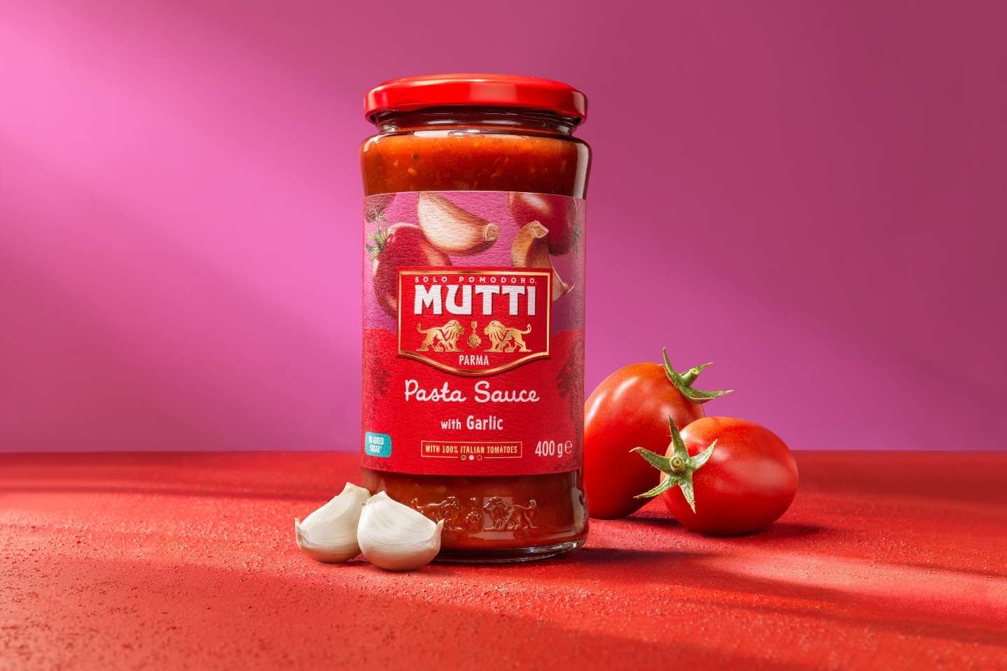 Mutti Pasta Sauce with Garlic -tomaattikastikepurkki valkosipulinkynsien ja tuoreiden tomaattien kanssa punaisella pinnalla.