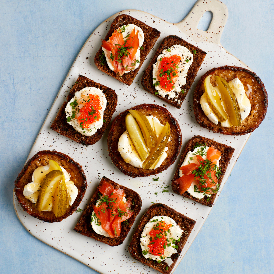 Herkullisia ruisleipäpaloja tarjoilulaudalla, päällä tuorejuustoa, lohta, mätiä ja suolakurkkua.