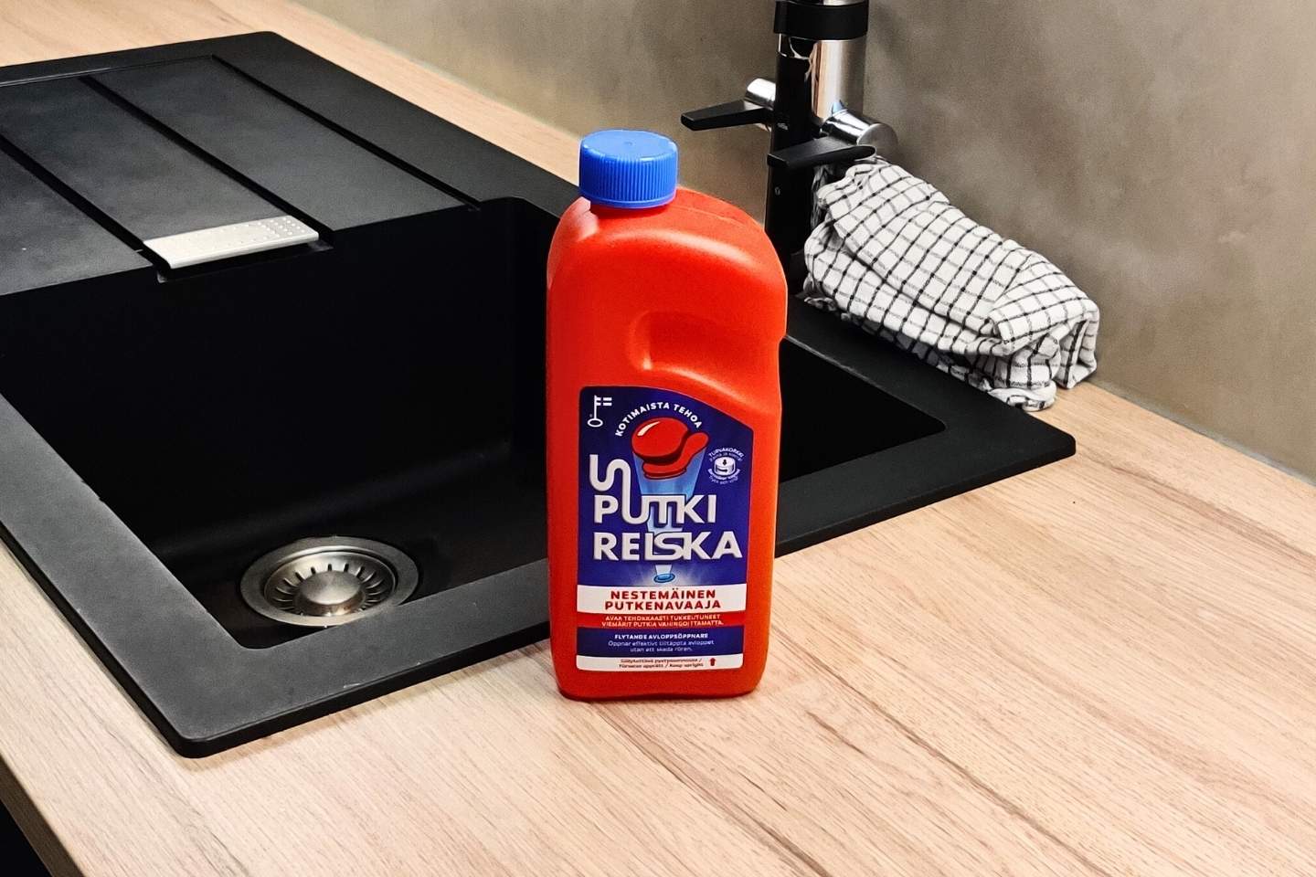 Punainen Putki Reiska -viemärinpuhdistusaine keittiötasolla mustan tiskialtaan vieressä, taustalla hana ja ruudullinen pyyhe.