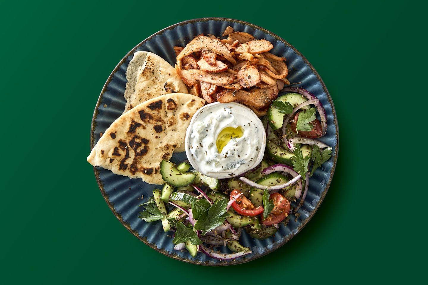 Sinisellä lautasella on shawarma-annos: mausteinen kana, pitaleipä, tzatziki ja tuore salaatti, jossa kurkkua, tomaattia ja punasipulia.