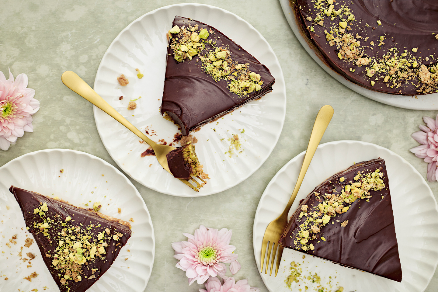 Herkullisia suklaakakkupaloja valkoisilla lautasilla, koristeltuna pistaasimurulla ja kultaisilla haarukoilla, vaaleanpunaisten kukkien ympäröimänä.