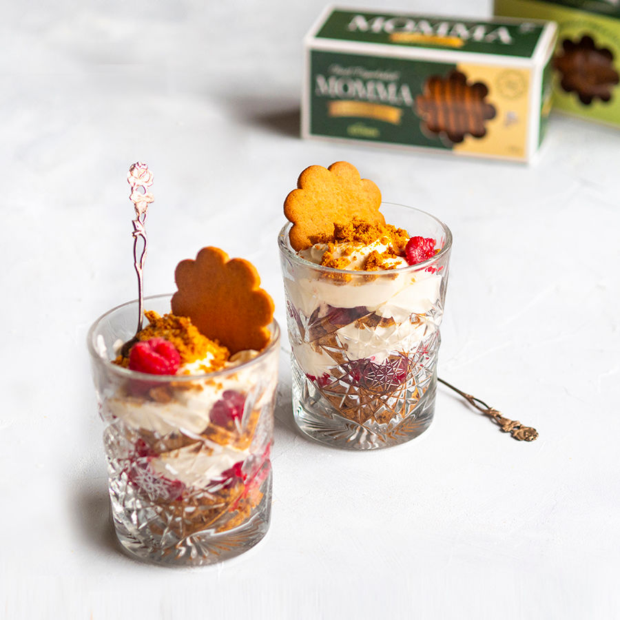 Kaksi koristeellista lasia täynnä herkullista jälkiruokaa, jossa kerroksia kermaa, vadelmia ja Mummia-keksejä koristeena.