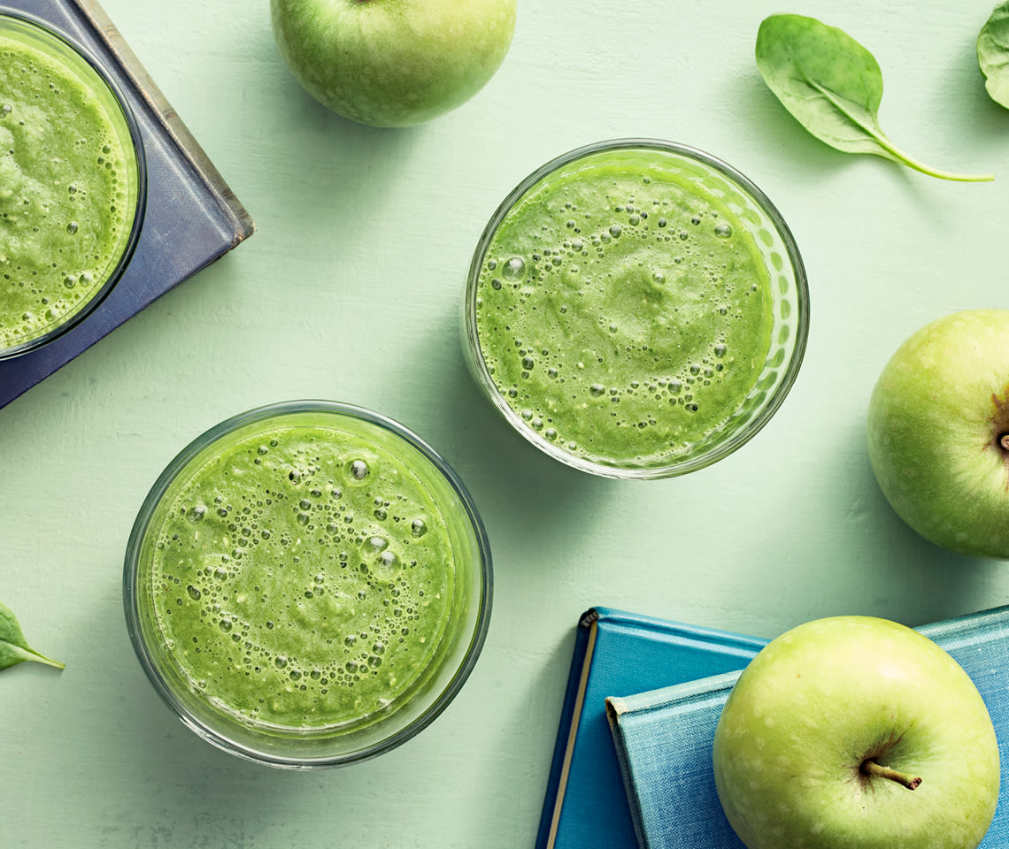 Kolme lasia vihreää smoothieta ylhäältä kuvattuna, ympärillä vihreitä omenoita ja pinaatinlehtiä vaalealla taustalla.
