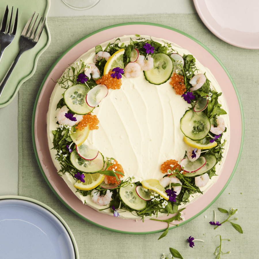 Pyöreä voileipäkakku vaaleanpunaisella lautasella, koristeltu katkaravuilla, kurkkuviipaleilla, mädillä ja syötävillä kukilla.