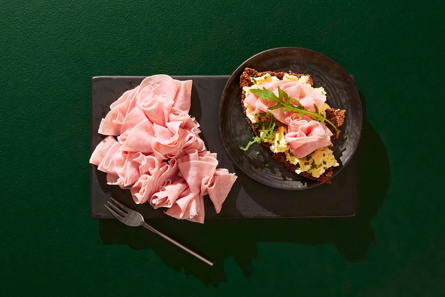 Herkullinen kinkkuannos: kasa ohutta kinkkuleikkelettä ja tumma leipäpala voilla, kinkulla ja yrteillä koristeltuna vihreällä taustalla.