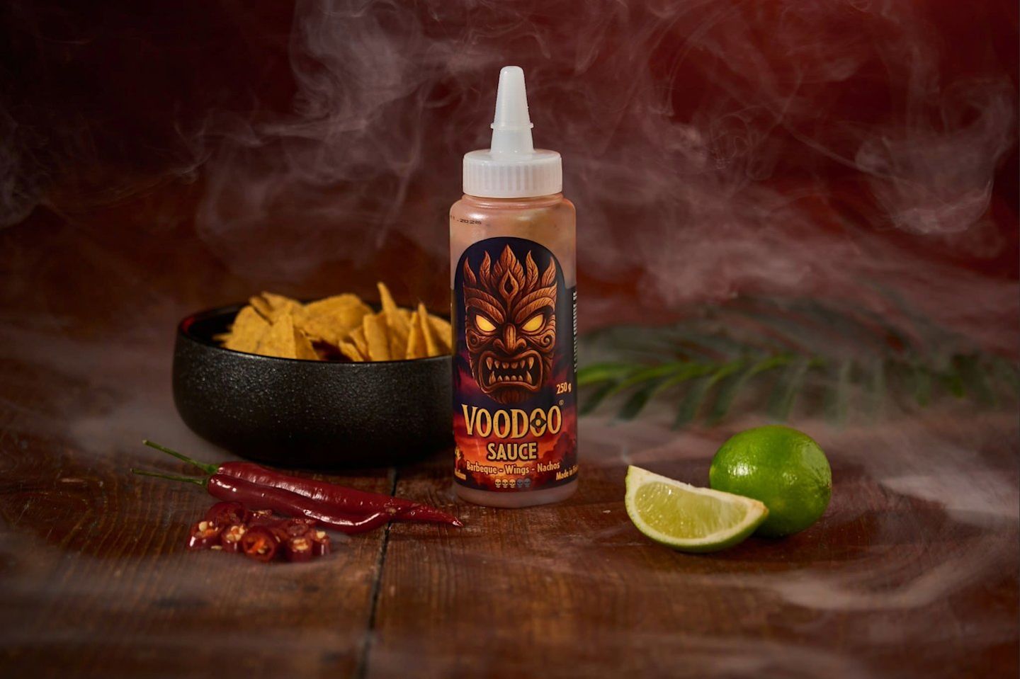 Voodoo Sauce -pullo puisella alustalla, ympärillä nachokulho, punaisia chilejä ja limettiä.