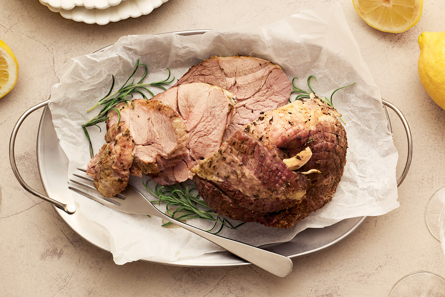 Mehevä karitsanpaisti leivinpaperilla, koristeltuna rosmariinilla ja sitruunaviipaleilla, tarjoiltuna vaalealla lautasella.