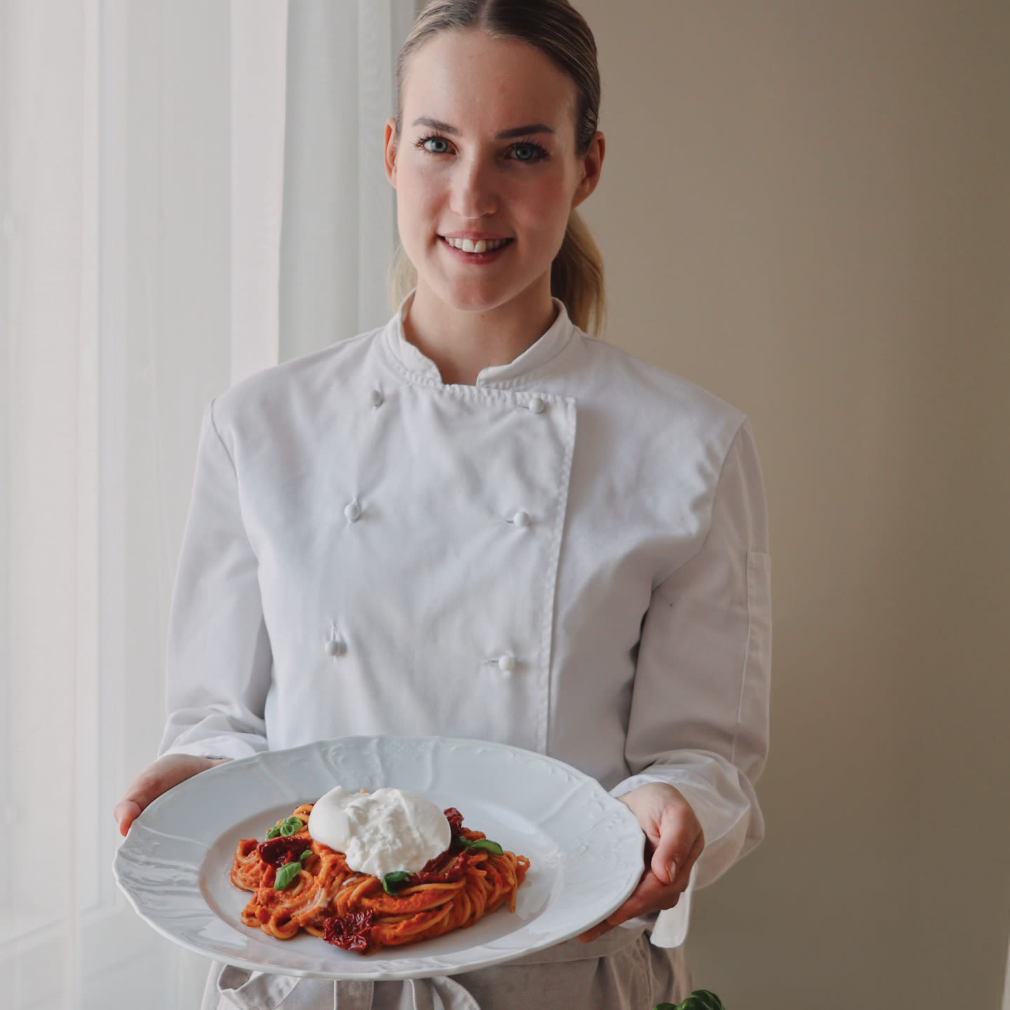 Hymyilevä kokki valkoisessa takissa esittelee annosta: pastaa tomaattikastikkeessa, päällä mozzarellaa ja basilikaa.