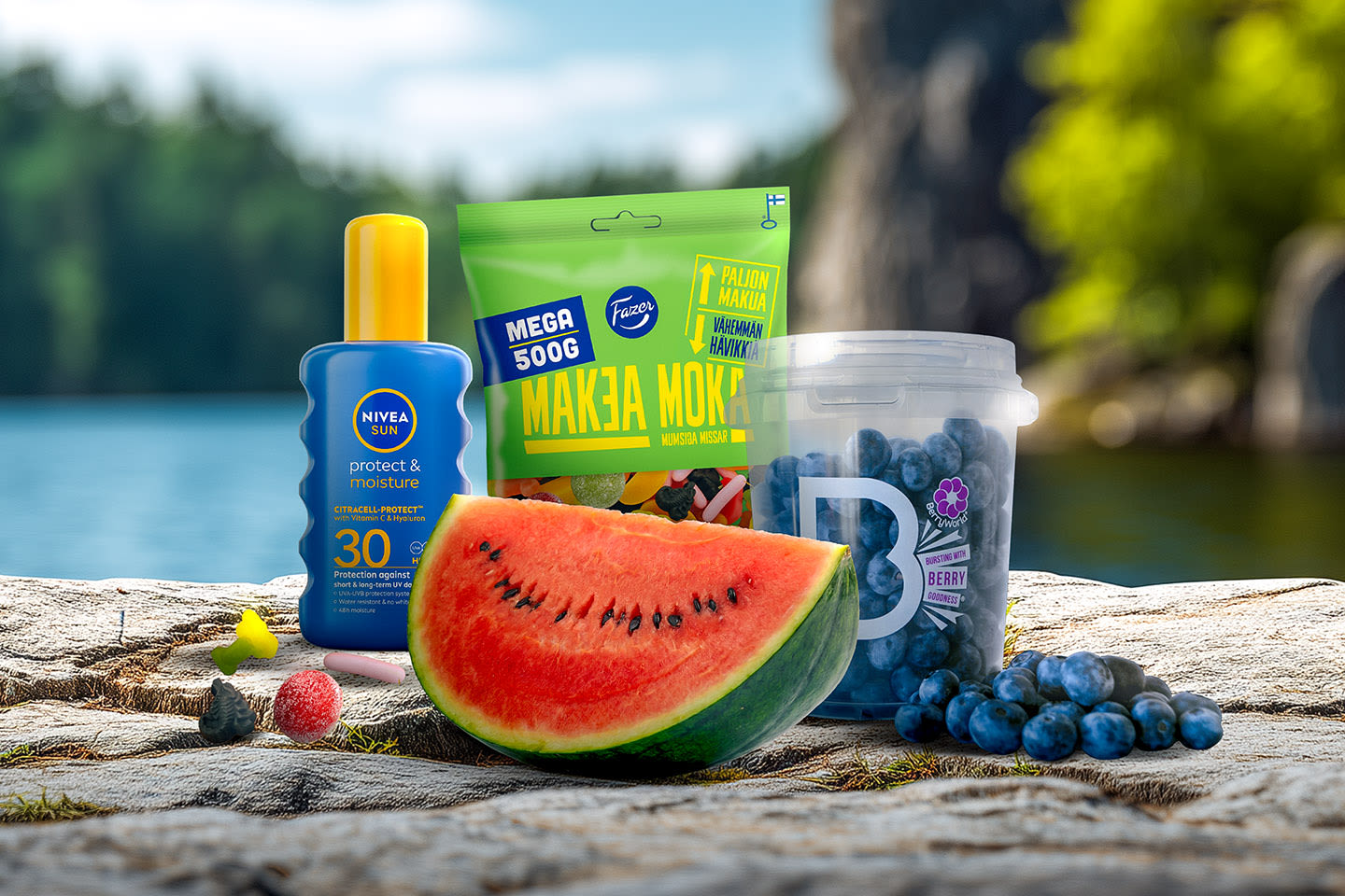 Kesäinen asetelma kalliolla: Nivea-aurinkovoide, Fazerin karkkipussi, vesimelonilohko ja mustikkarasia järvimaiseman edessä.
