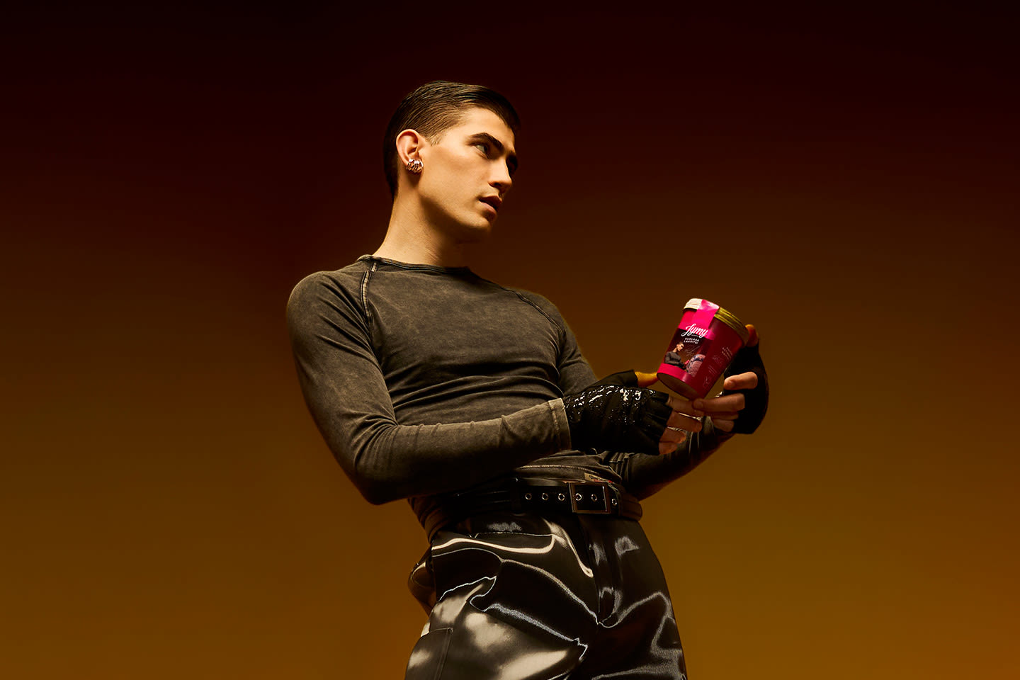 Henkilö tummassa paidassa ja nahkahanskoissa pitää kädessään kirkkaanpunaista tuotepurkkia lämpimässä, ruskeansävyisessä valaistuksessa.