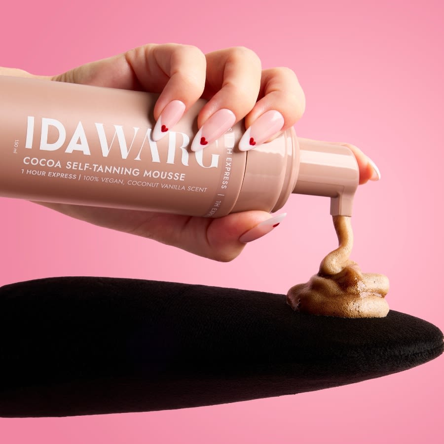 Käsi pumppaa Ida Warg Cocoa Self-Tanning Mousse -itseruskettavaa vaahtoa mustalle levityskintaalle, vaaleanpunainen tausta.