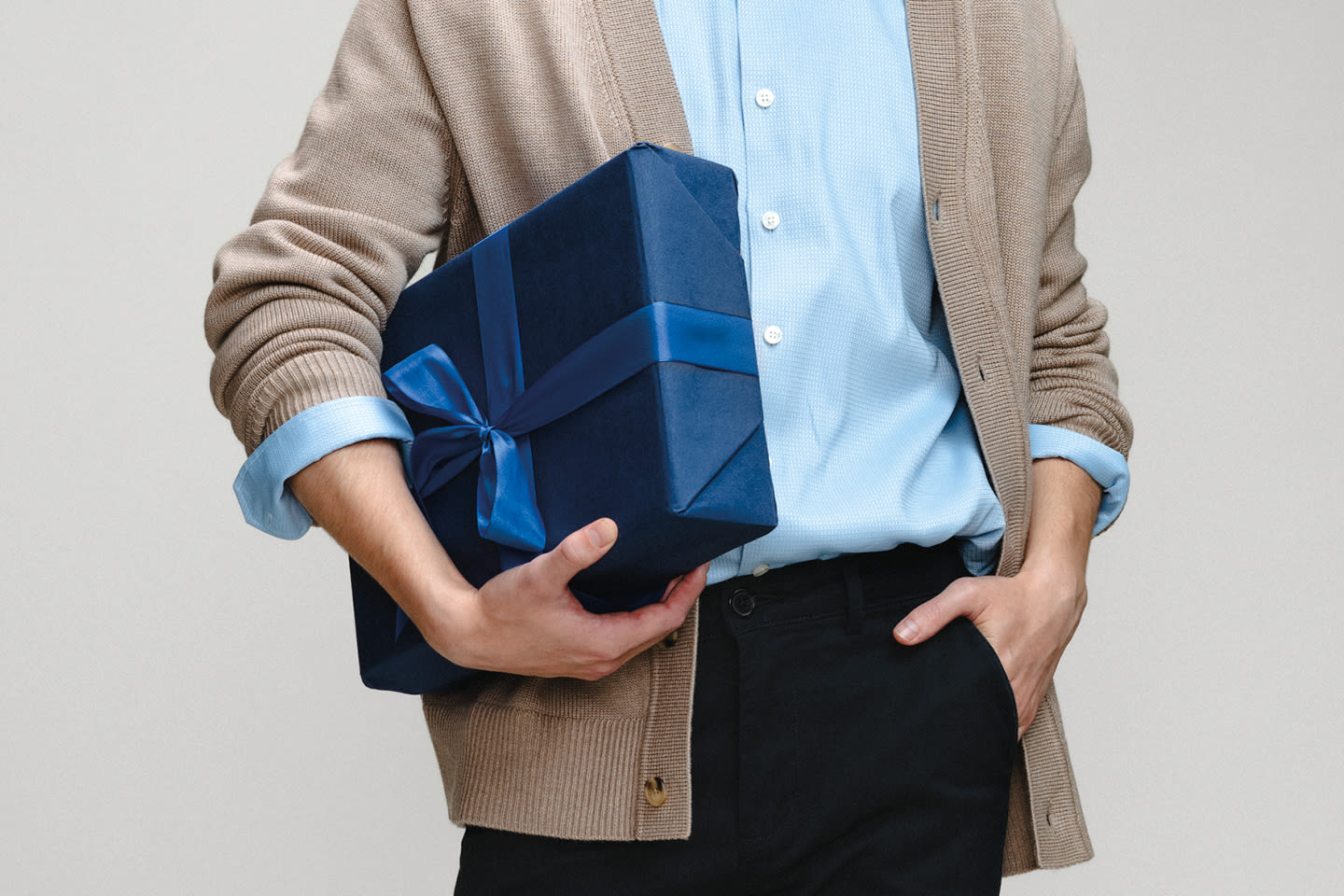 Henkilö beigessä neuletakissa ja vaaleansinisessä paidassa pitää kädessään tyylikkäästi pakattua sinistä lahjapakettia.