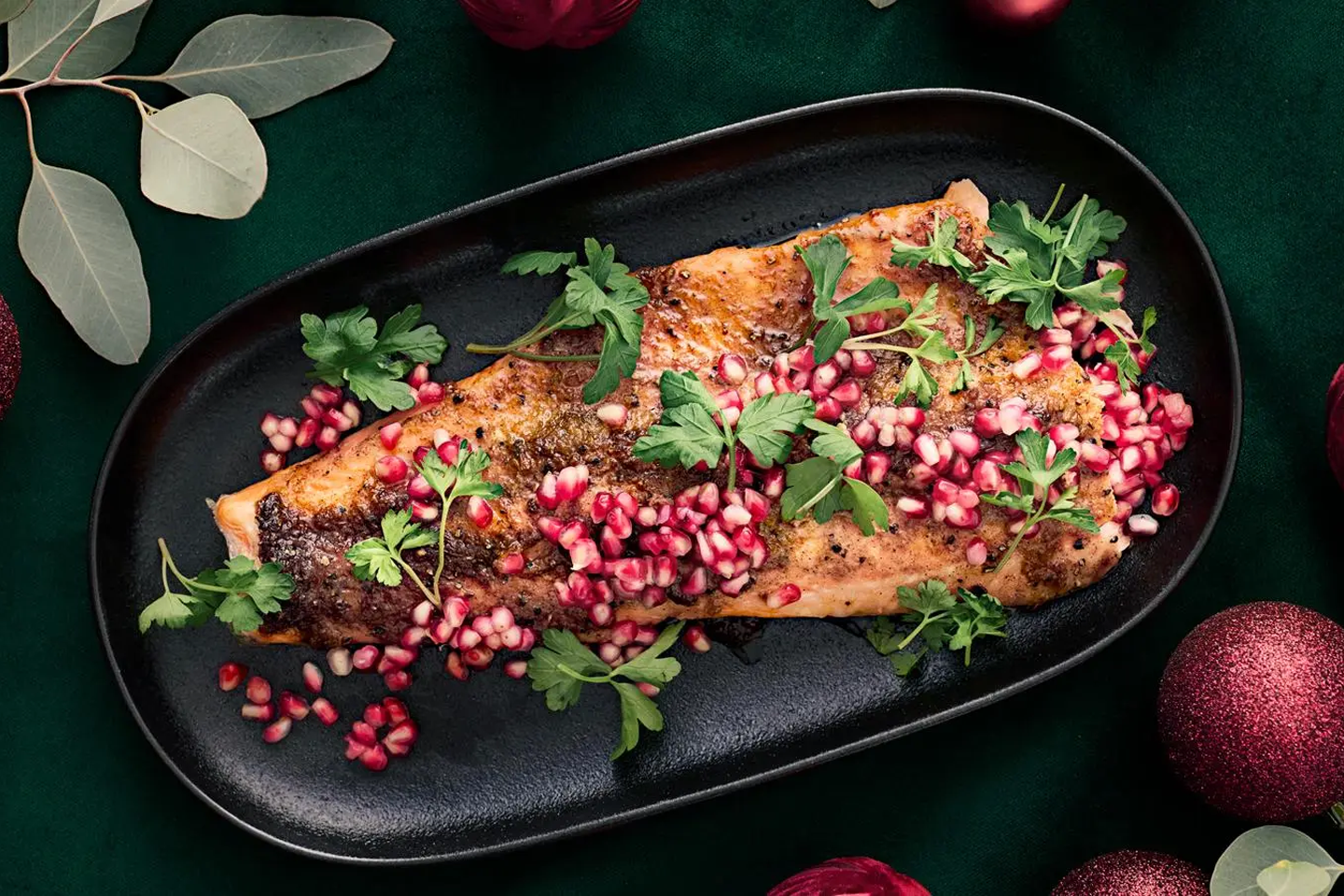Mehevä paistettu kala mustalla lautasella, koristeena granaattiomenan siemeniä ja persiljaa, vihreällä taustalla joulukoristeet.