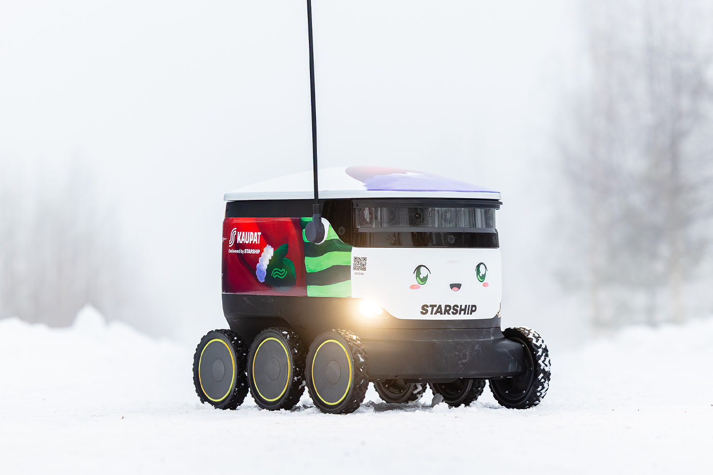 Söpö Starship-kuljetusrobotti liikkuu lumisessa maisemassa, etuvalojen loistaessa ja S-kaupat-logon koristamana.