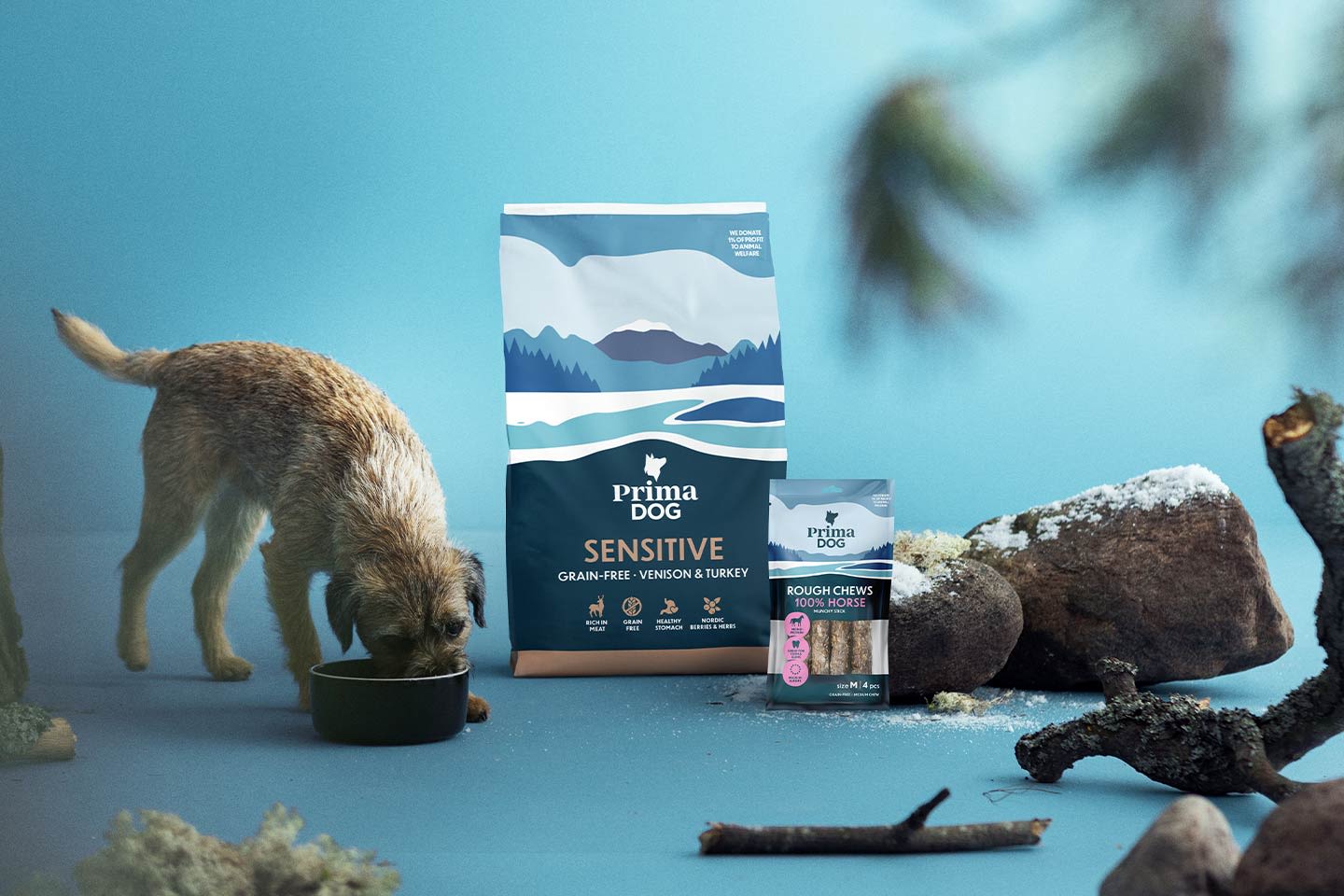 Pieni ruskea koira syö kupista Prima Dog Sensitive -ruokapussin ja Rough Chews -purutikkupakkauksen vieressä sinisellä taustalla.
